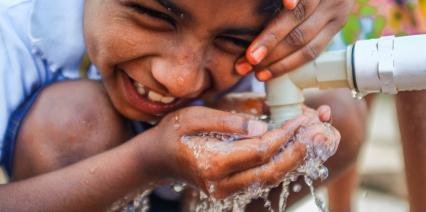 Indian boy smiling and drinking water from a communal faucet.