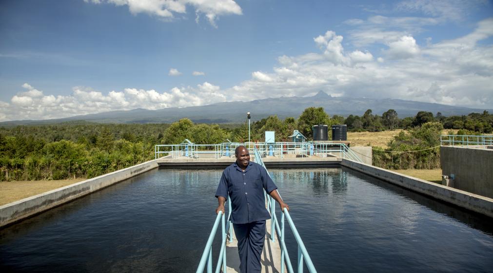 A man walking on a narrow walkway above a water treatment facility with mountains and greenery in the background.